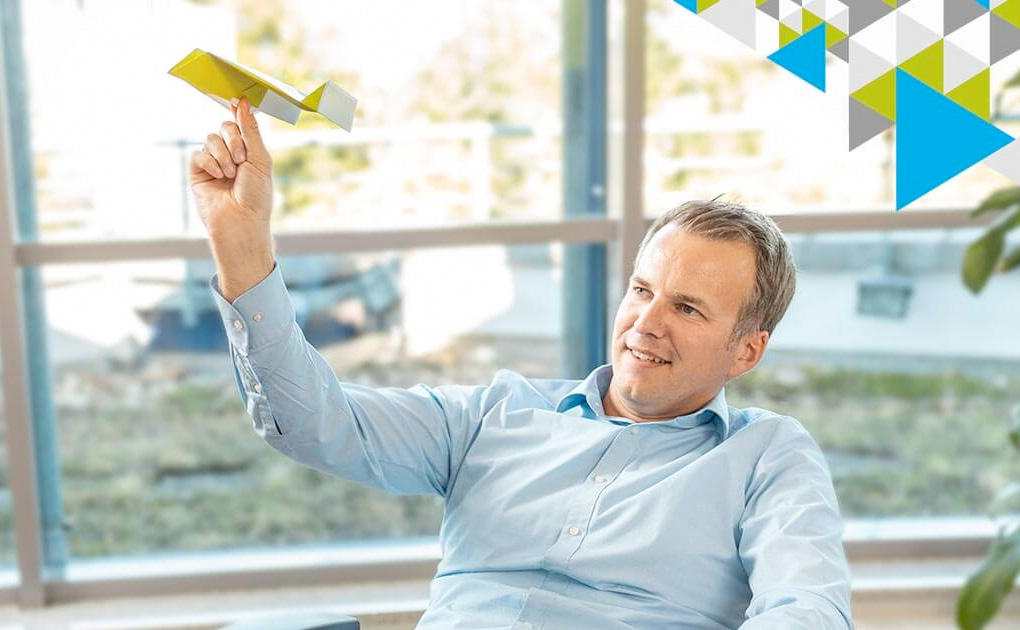 Mann in blauem Hemd hält Papierflieger in der Hand vor großen Fenstern mit Blick ins Freie