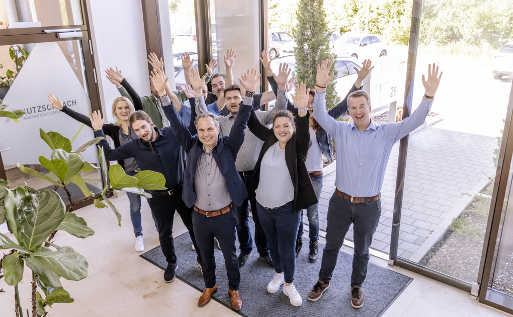 In a bright entrance area of ​​an office building, a cheerful group of several people raise their hands in celebration. They appear relaxed and inviting, which indicates a positive company culture.