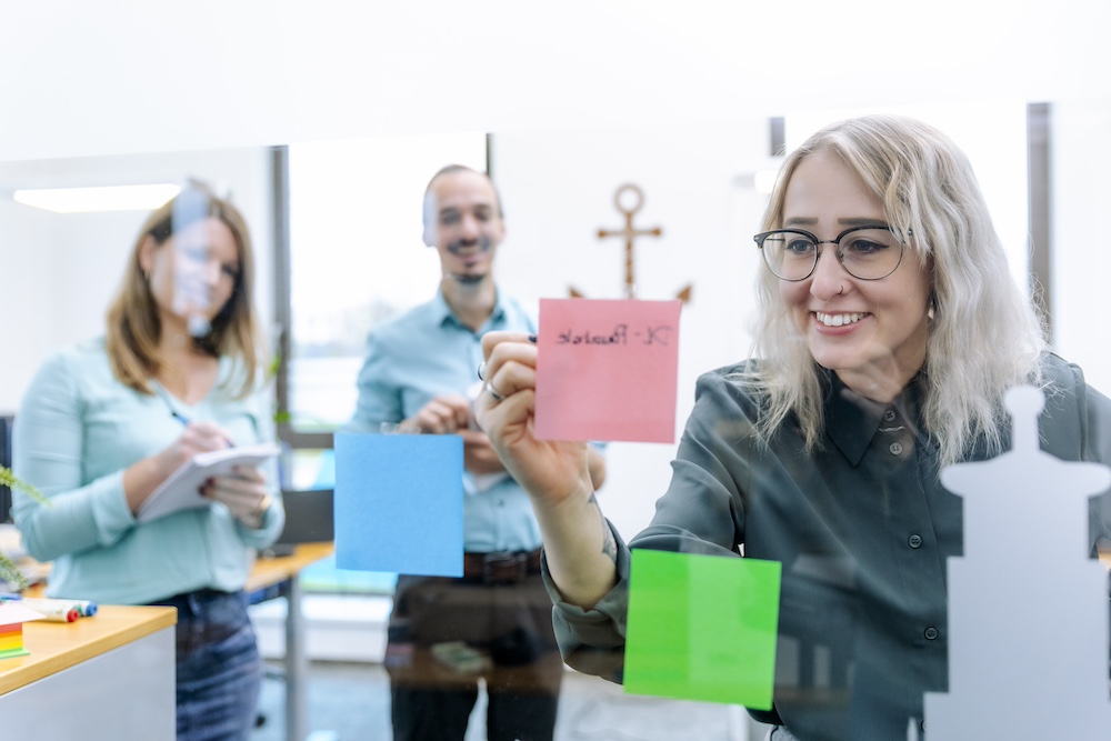 A woman with glasses presents colored sticky notes on a glass wall, a man and a woman in the background also look at notes. You are in a brainstorming meeting and seem engaged and cooperative.