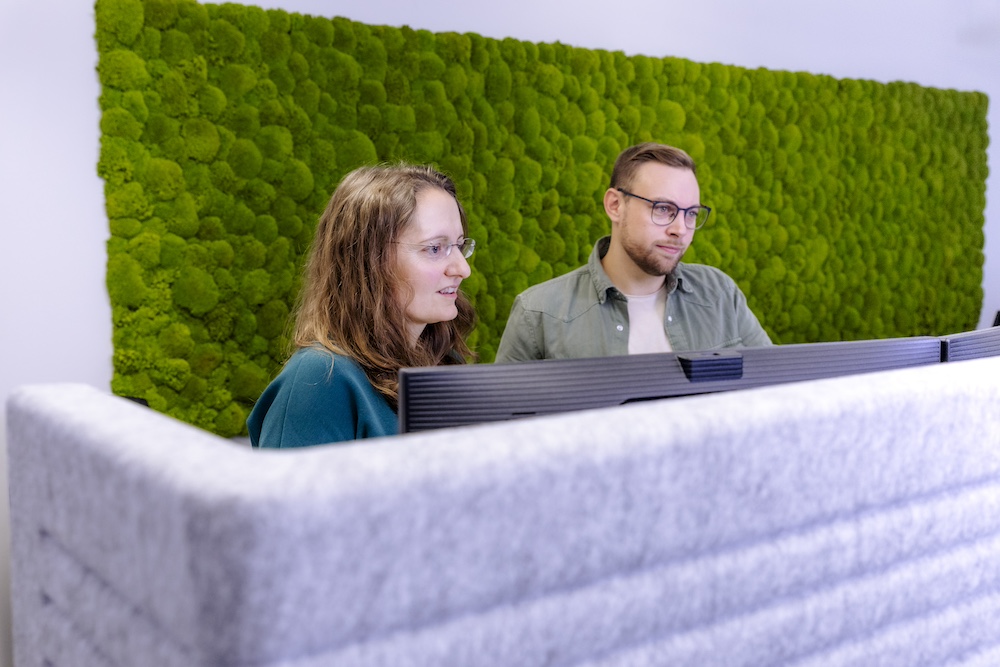 Two office workers, a woman and a man, work together on a computer in an office environment. Behind them is a striking green wall made of moss, creating a natural and calming background. They seem focused and committed to their task.