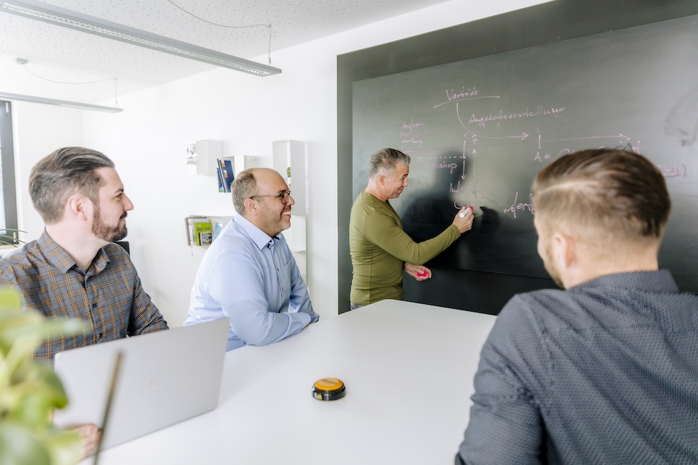 Four men in a bright office during a brainstorming session. One of the men stands at a blackboard and writes while the others watch and listen. The board is filled with notes and diagrams, indicating a collaborative work environment and teamwork.