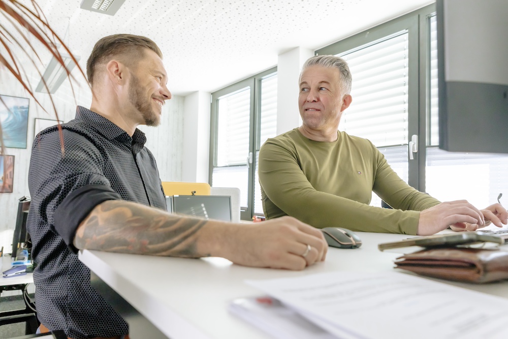 Zwei Männer im Gespräch am Arbeitsplatz, einer mit Tattoos und lächelnd, der andere sitzend und schreibend, beide in einer hellen, modernen Büroumgebung. Sie scheinen in eine angenehme Unterhaltung vertieft zu sein.