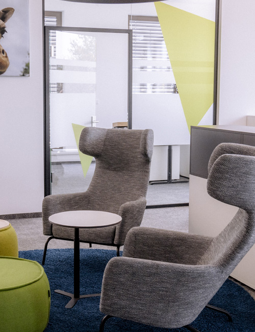 A modernly furnished office with two high gray armchairs, a round side table and color-coordinated green stools. The glass wall in the background is designed with geometric yellow and green foils that add an artistic touch and brighten the room.