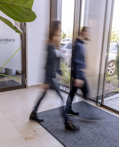 Two blurred people walking through a glass door in an office building, suggesting movement. The scene conveys a dynamic work environment.