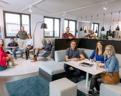 A team of nine employees in a modern office, spread out on sofas and armchairs, some sitting and others standing. They appear to be interacting in a relaxed business atmosphere, perhaps during a break or in an informal meeting.