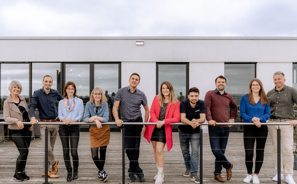 Ein Gruppenfoto von zehn Personen, die lächelnd in einer Reihe am Geländer einer Außenterrasse stehen, mit einem Gebäude im Hintergrund. Sie scheinen in entspannter Geschäftskleidung gekleidet zu sein und posieren für ein Teamfoto.