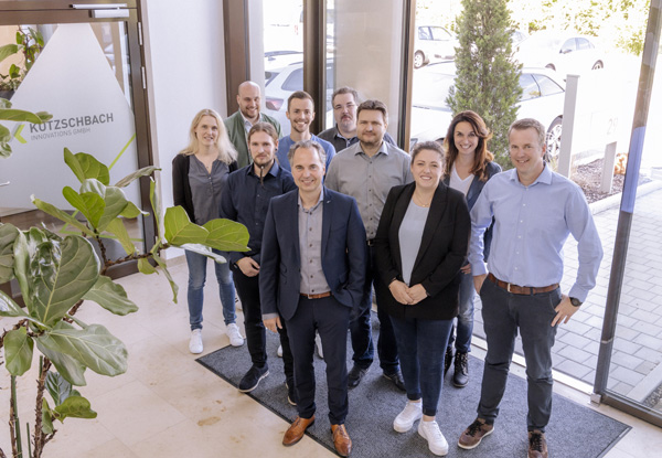 Group photo of the Kutzschbach team in Augsburg. The people stand in the entrance area and pose smiling. The company logo is visible in the background on the glass door.