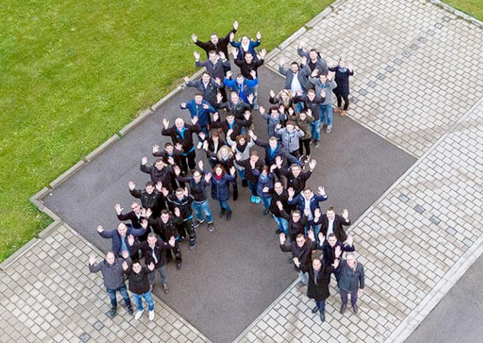 Aerial view of a large group of people standing on a lawn shaped in the shape of the letter K and waving upwards at the camera.