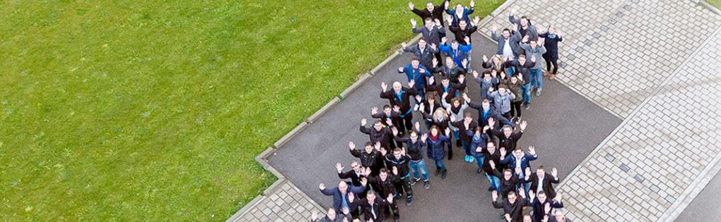 Aerial view of a large group of people standing on a lawn shaped in the shape of the letter K and waving upwards at the camera.