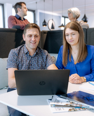 Ein Mann und eine Frau sitzen zusammen und blicken auf einen Laptop-Bildschirm, im Hintergrund sind Büromöbel und weitere Kollegen zu sehen.
