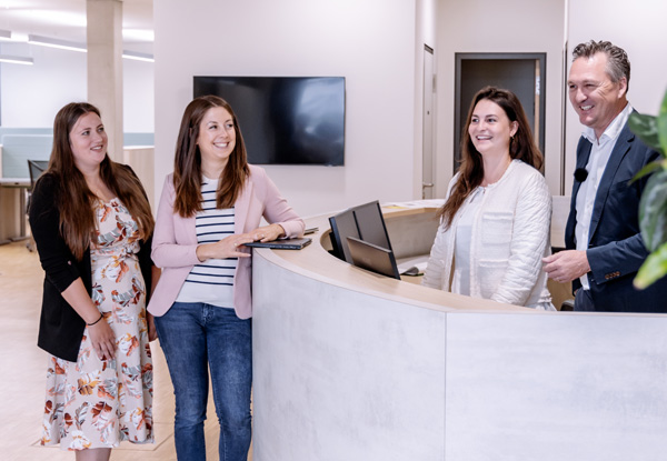 Three women and a man stand smiling behind a reception desk in a bright office building.