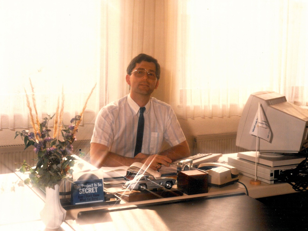 Ein Mann in einem Büro sitzt an einem Schreibtisch neben einem Blumenstrauß und vor einem alten Computer, mit einem humorvollen Schild, das "This project is so secret" sagt. Das Bild ist aus den 80iger Jahren.