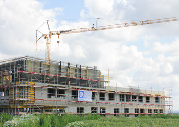Construction site of a multi-story building with a crane and scaffolding under a cloudy sky surrounded by green vegetation.