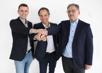 Three men in suits stand next to each other and put their hands together in a gesture of teamwork, smiling in front of a bright window.