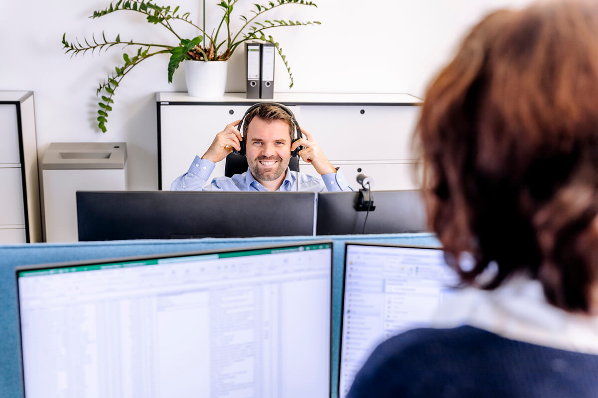 Ein Mann in einem blauen Hemd setzt gerade ein Headset auf und lächelt, während er an einem Computerarbeitsplatz sitzt.