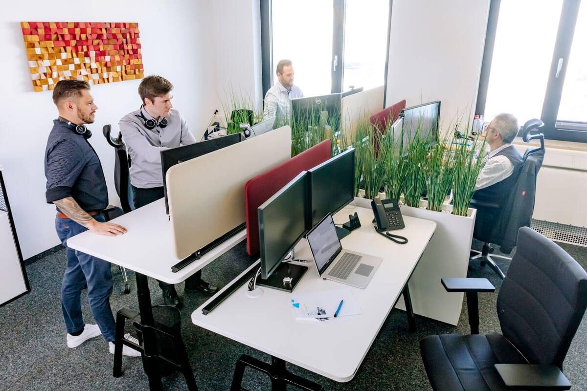 A modern office with four people at standing desks working intently on their computers. The office is bright and decorated with abstract wall art and plants as a room divider, creating a creative and lively working atmosphere. People are dressed casually and professionally, indicating a relaxed but productive work environment.