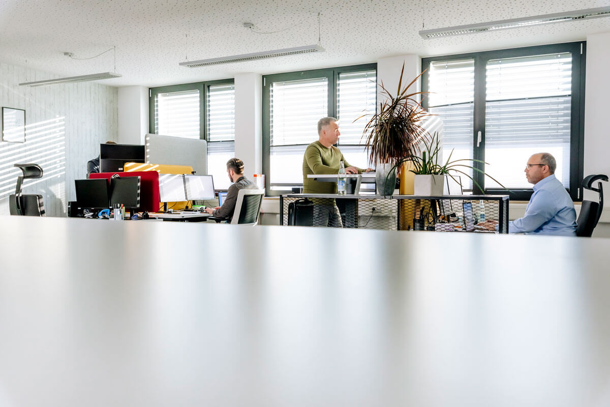 Ein offenes Büro mit vier Männern, die verschiedene Tätigkeiten ausführen. Der Raum ist hell mit großen Fenstern und ist mit Büropflanzen ausgestattet, die eine lebendige Arbeitsatmosphäre schaffen.