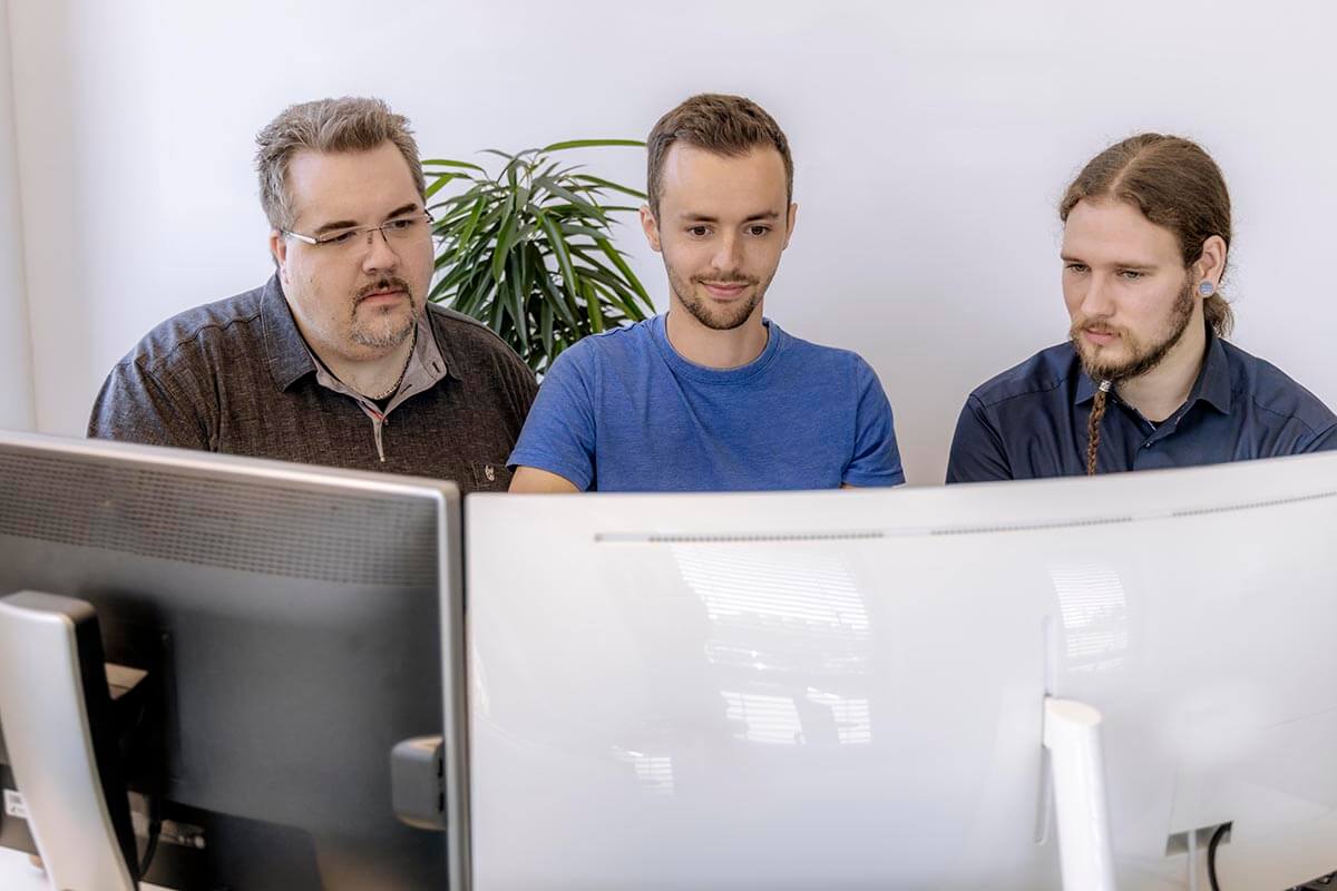 Three men look over the monitors at their workstations in a bright office. You smile warmly at the camera and represent a diverse team in a collaborative work environment. A houseplant can be seen in the background, which also creates a pleasant office atmosphere.