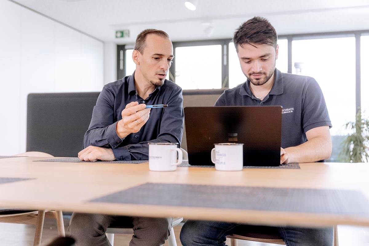 Zwei Kollegen, einer in einem dunkelblauen Hemd und der andere in einem dunklen Polo-Shirt, betrachten gemeinsam etwas auf einem Laptop auf einem Schreibtisch. Sie scheinen in eine konstruktive Diskussion vertieft zu sein.