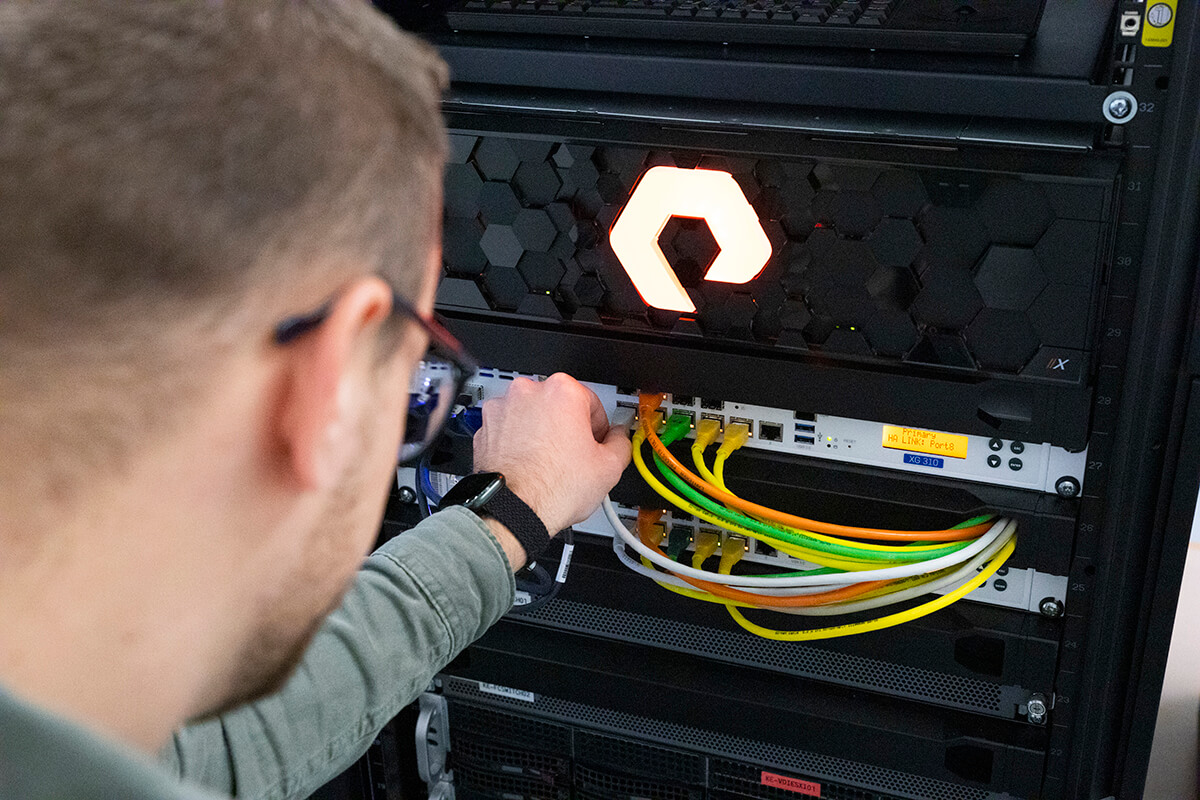 Ein Techniker arbeitet konzentriert an einem Serverschrank mit einer beleuchteten Plexiglasfront in Form eines Logos. Man sieht Netzwerkkabel, die sorgfältig verbunden werden, was auf eine Wartungs- oder Einrichtungstätigkeit im Bereich der Informationstechnologie hindeutet.
