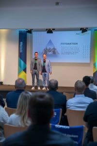 Zwei Geschäftsführer stehen auf Bühne beim Kutzschbach Partnertag 2025 vor Publikum, Präsentation mit 'Fotoaktion' auf Leinwand