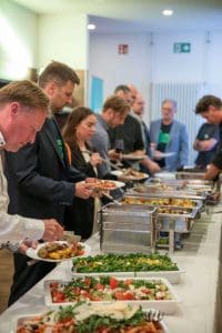 Teilnehmer des Kutzschbach Partnertag 2025 bedienen sich an einem Grillbuffet mit verschiedenen warmen und kalten Speisen in einem Veranstaltungsraum