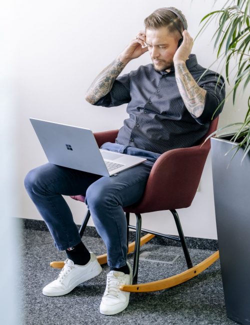 A concentrated man with tattoos sits in a modern rocking chair, a laptop on his knees, touching his head as if thinking or seeking a solution. He is dressed casually and is in a bright corner of the office next to a large potted plant that creates a calming atmosphere.