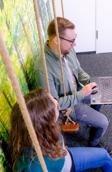 Ein Mann und eine Frau sitzen auf Schaukeln in einem Büro, umgeben von Wandtapeten mit Waldmotiv. Sie sind in eine Laptop-Arbeit vertieft, was eine kreative und entspannte Arbeitsumgebung suggeriert.