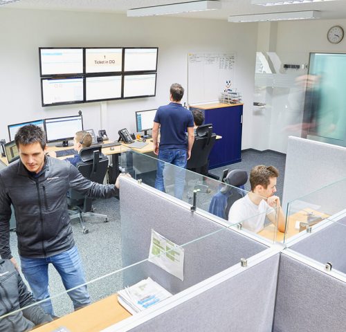 Vogelperspektive auf ein belebtes Büro mit Trennwänden, an dem Mitarbeiter an Computern arbeiten und auf große Monitore an der Wand schauen.