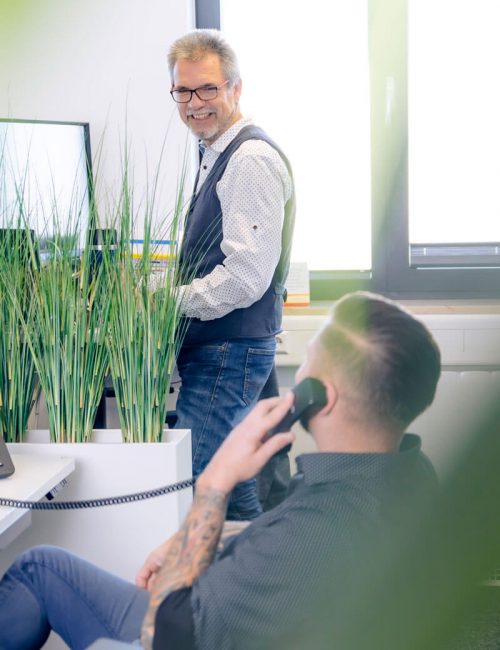 Ein lächelnder Mann schaut über einen Monitor hinweg auf einen sitzenden Kollegen. Die Szene ist durch Pflanzen im Vordergrund teils verdeckt, was eine lebendige und freundliche Arbeitsatmosphäre suggeriert. Helle und natürliche Farbtöne dominieren den Raum.