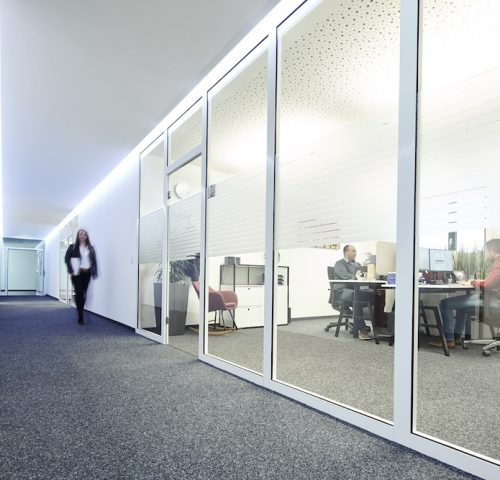 View of a bright hallway in a modern office building, with blurred people working in offices and one person walking down the hallway.