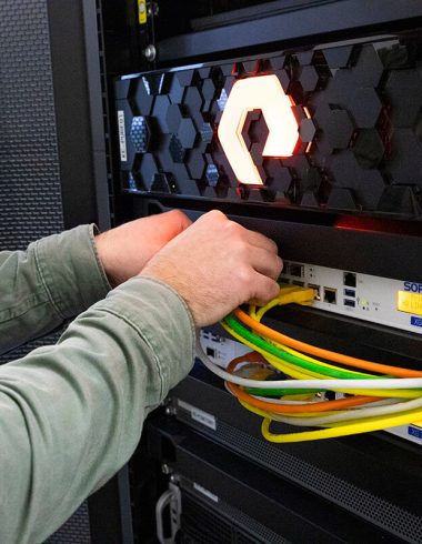 A technician works concentratedly on a server cabinet with an illuminated Plexiglas front in the shape of a logo. Network cables can be seen being carefully connected, indicating information technology maintenance or setup activity.