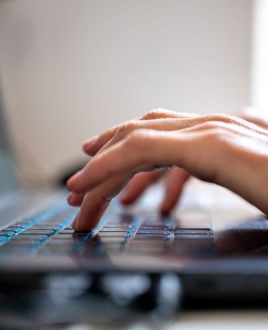 Eine Nahaufnahme einer Hand, die auf einer Laptop-Tastatur tippt, mit einem Fokus auf die Finger, die gerade eine Taste drücken, was auf eine aktive Texteingabe oder Programmierung hinweist.